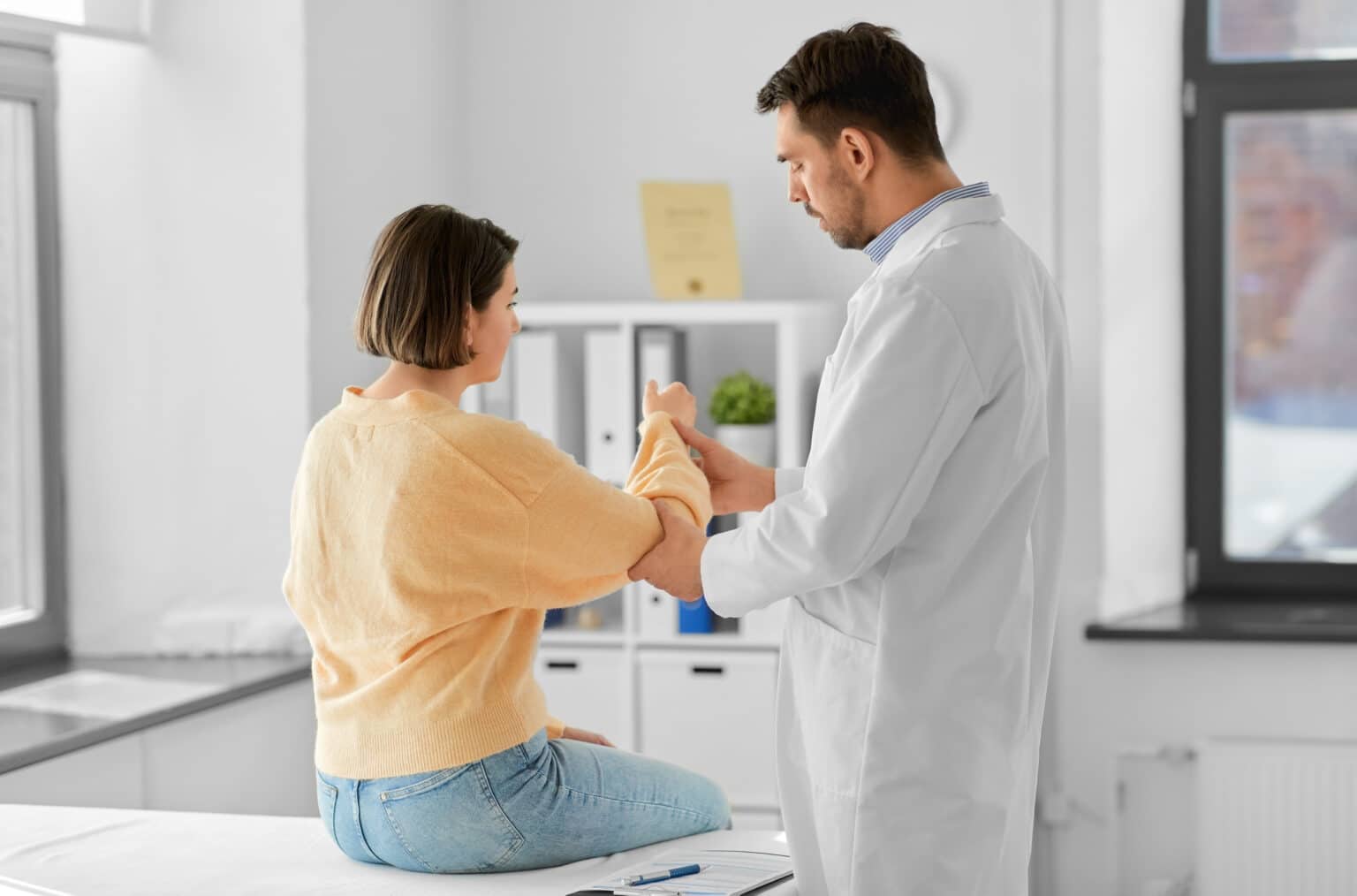 medicine, healthcare and people concept - male doctor with clipboard talking to woman showing her sore arm patient at hospital